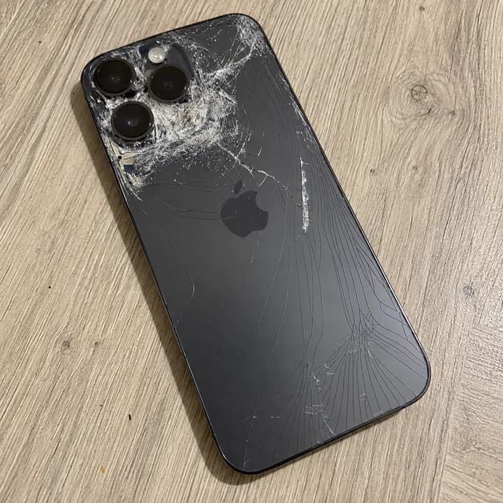 Grafitni iPhone z močno razbitim hrbtnim steklom na leseni površini.
