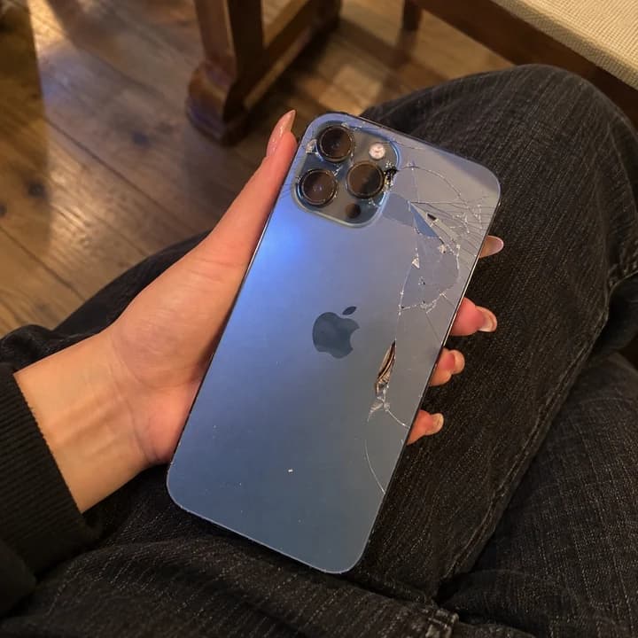 Moder iPhone v roki z razpokami in manjkajočimi koščki hrbtnega stekla.